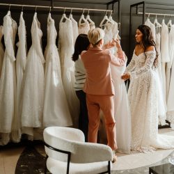 A woman stands and smiles while listening to two women standing across from her holding a dress, in a bridal shop, during wedding preparations